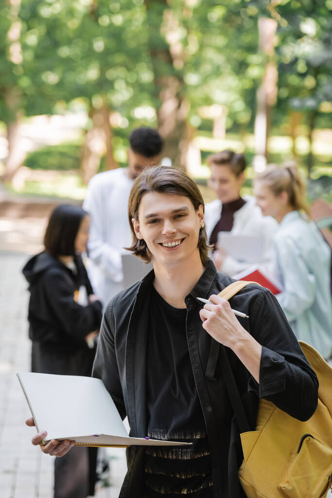Usmívající se student s notebookem a batohem při pohledu do kamery v blízkosti rozmazaných přátel v parku  - Fotografie, Obrázek