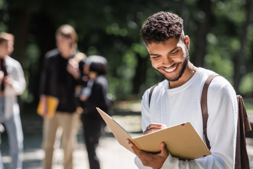 Sırt çantalı gülümseyen Afrikalı Amerikalı öğrenci açık havada deftere yazıyor.  - Fotoğraf, Görsel