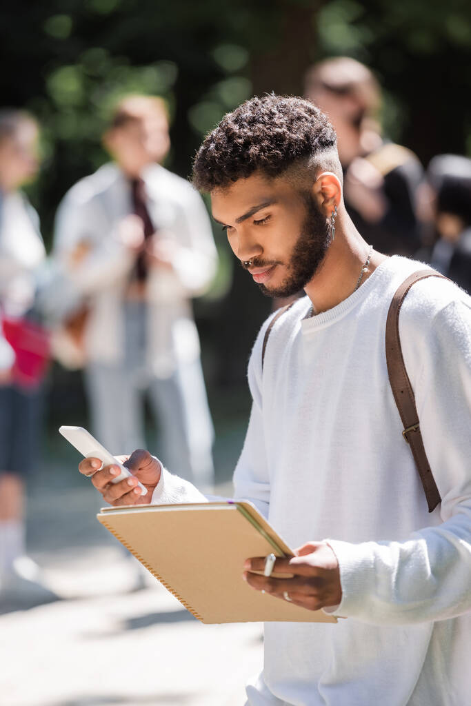 Estudiante afroamericano sosteniendo teléfono inteligente y portátil en el parque  - Foto, Imagen