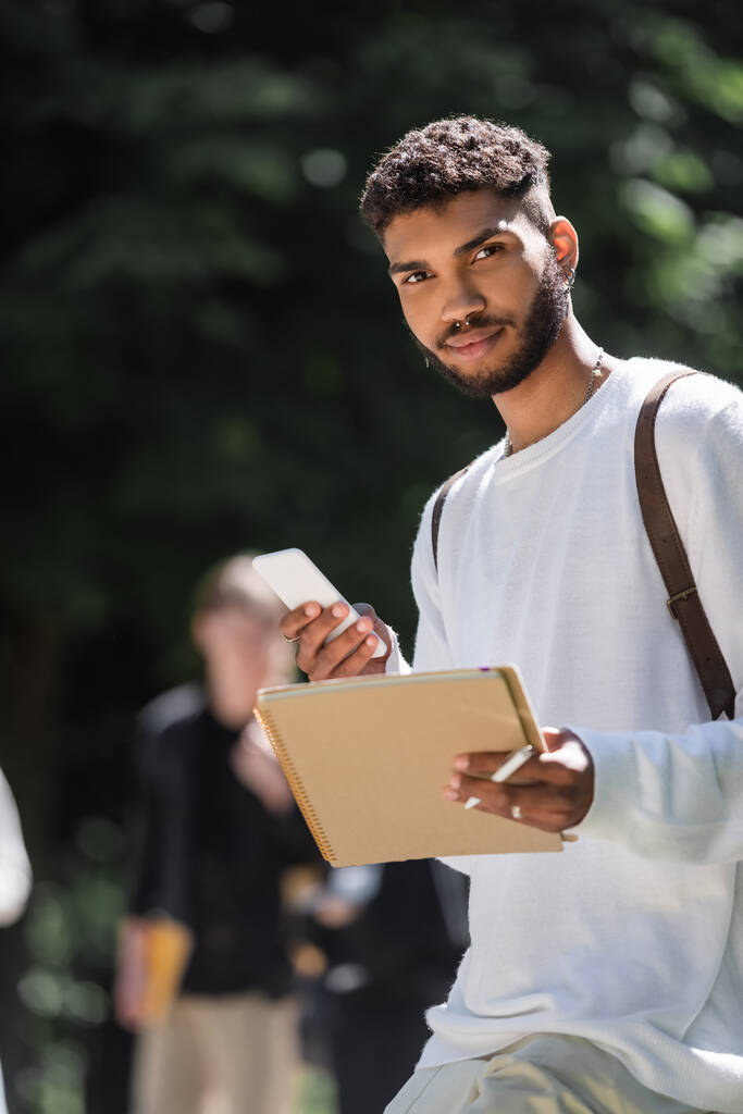Mladý africký americký student držící smartphone a notebook v parku  - Fotografie, Obrázek