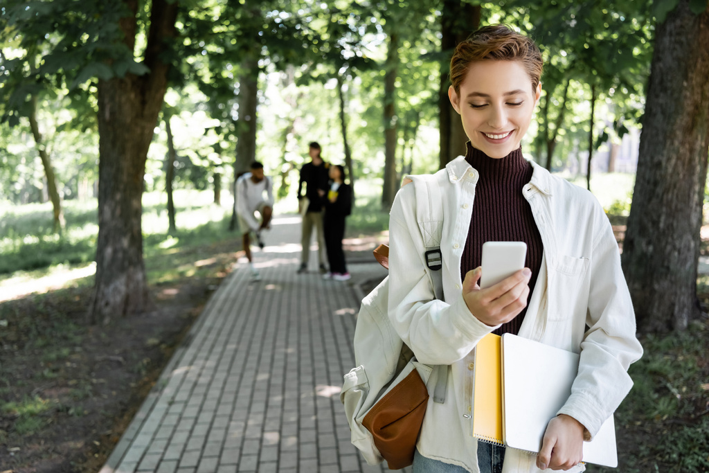 Усміхнений студент з ноутбуком за допомогою смартфона біля розмитих друзів в літньому парку
  - Фото, зображення
