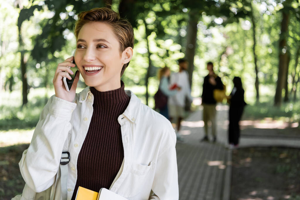 Pozitivní student s batohem mluví na mobilu v rozmazaném parku  - Fotografie, Obrázek