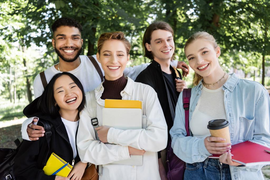 Cheerful interracial students with notebooks looking at camera in summer park  - Foto, Imagem