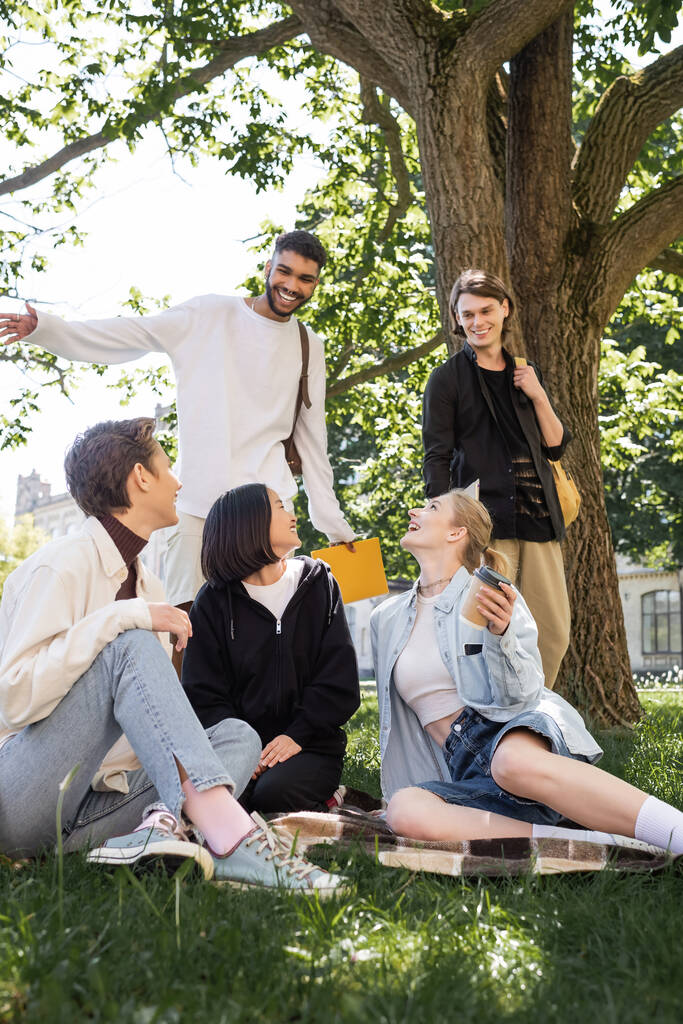 Позитивные межрасовые студенты с рюкзаками смотрят на друзей на одеяле в парке  - Фото, изображение