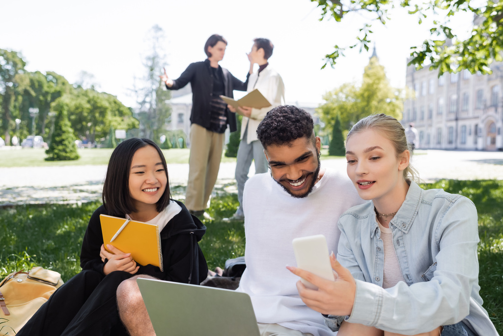 Student gazdaság okostelefon közelében mosolygós multikulturális diákok laptop és notebook füvön a parkban  - Fotó, kép