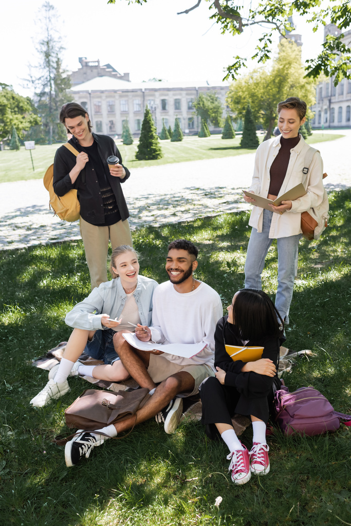 Веселые межрасовые студенты с книжками, проводящие время на траве в парке  - Фото, изображение