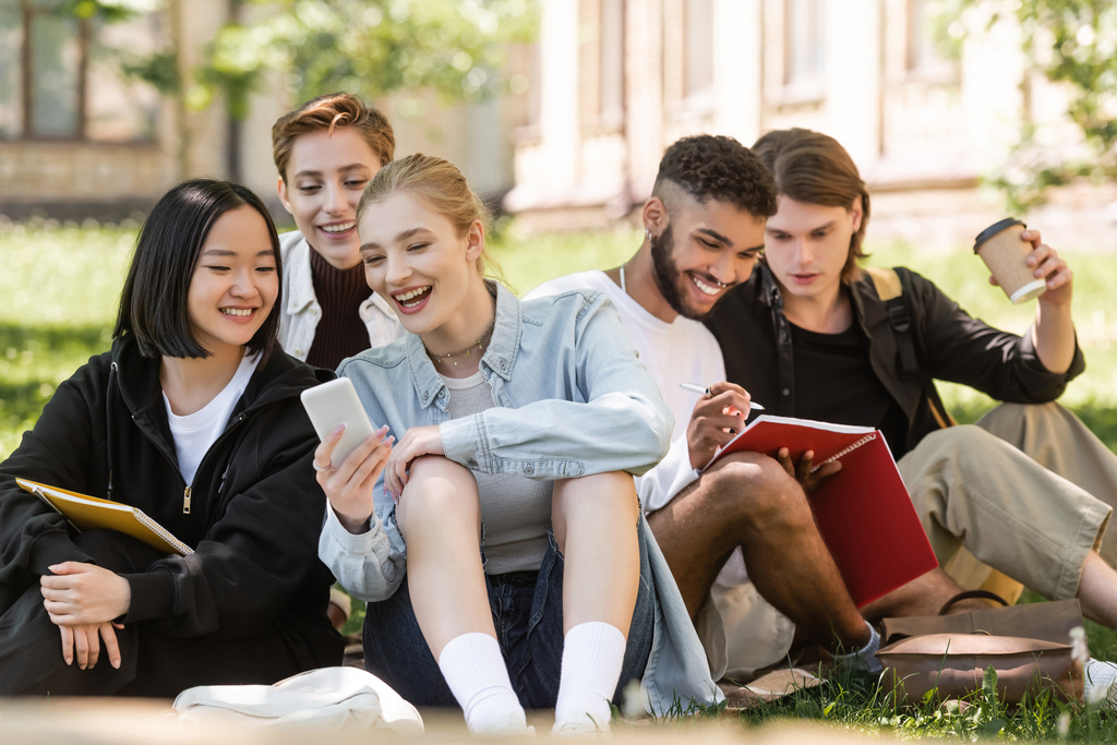 Smiling interracial students looking at smartphone near friends with notebook on lawn in park  - Foto, Imagen