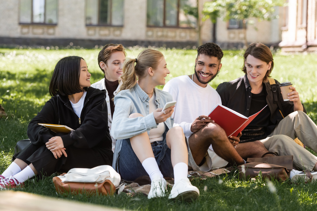Мультикультурные студенты со смартфоном и копировальными книгами проводят время на траве в парке  - Фото, изображение