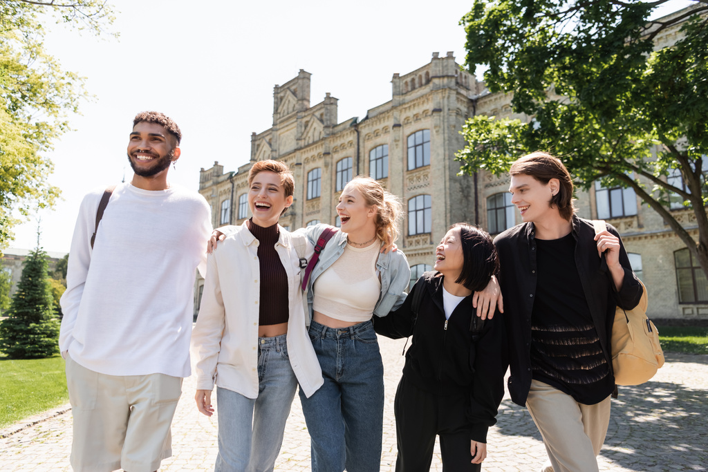 Gruppo di studenti multiculturali sorridenti che si abbracciano mentre camminano vicino all'università all'aperto  - Foto, immagini