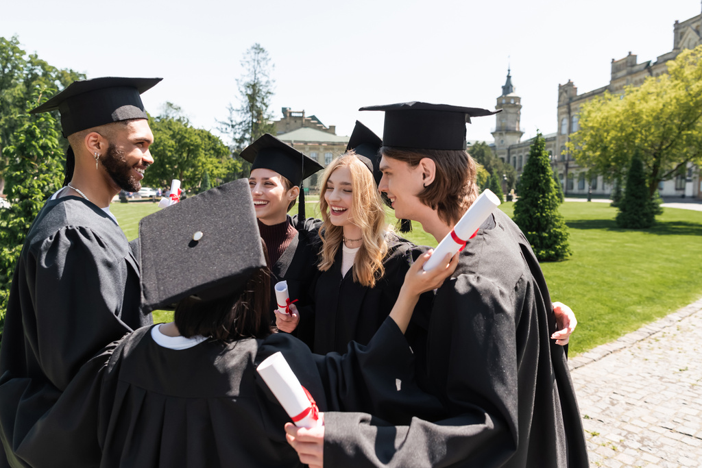 Усміхнені міжрасові холостяки в сукнях тримають дипломи і обіймаються в парку
  - Фото, зображення
