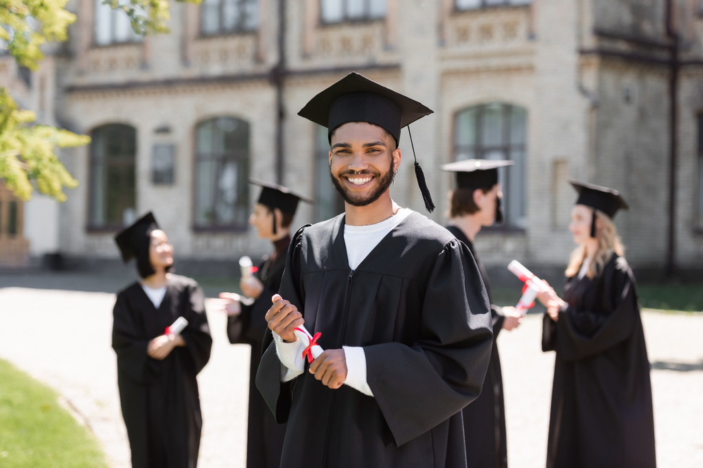 Licenciatura afroamericana en gorra y vestido con diploma cerca de amigos borrosos y la universidad al aire libre  - Foto, imagen