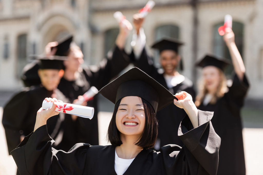 Sonriente soltero asiático en la tapa y vestido de la celebración de diploma cerca borrosa amigos al aire libre  - Foto, imagen