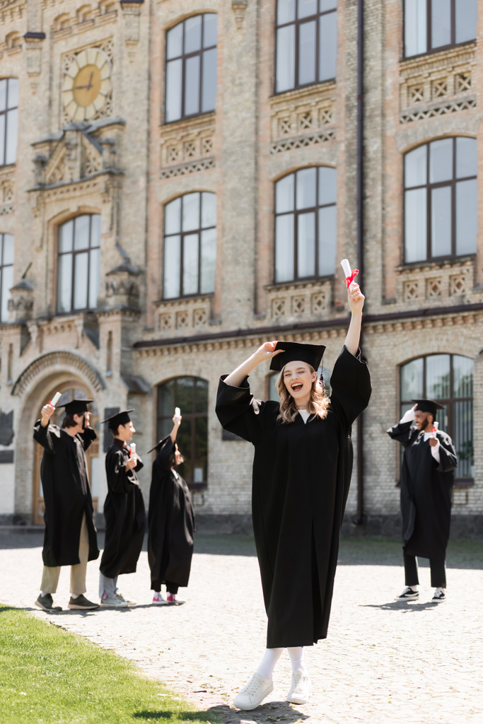 Excited bachelor holding diploma near blurred multiethnic friends and university outdoors  - Foto, Imagem