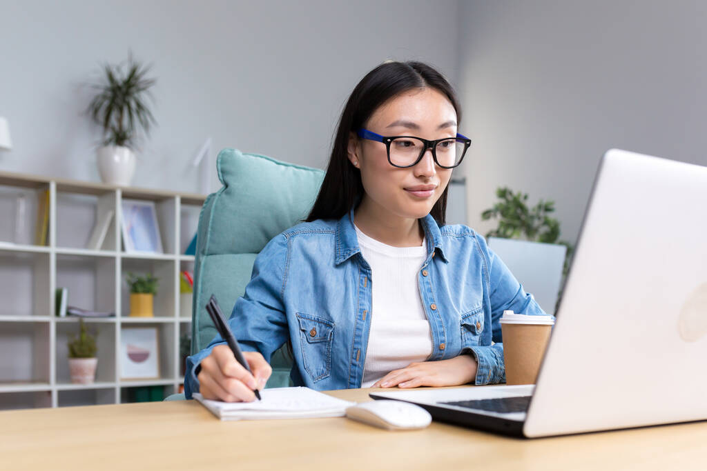 Fiatal ázsiai nő tanul az interneten, miközben ül az irodában, jegyzeteket ír a jegyzetfüzetbe, laptopot használ a tanuláshoz. - Fotó, kép