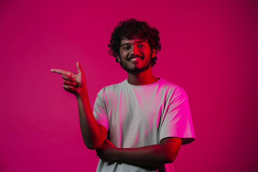 Молодий середній східний чоловік посміхається і вказує палець убік ізольовано над рожевою стіною
 - Фото, зображення