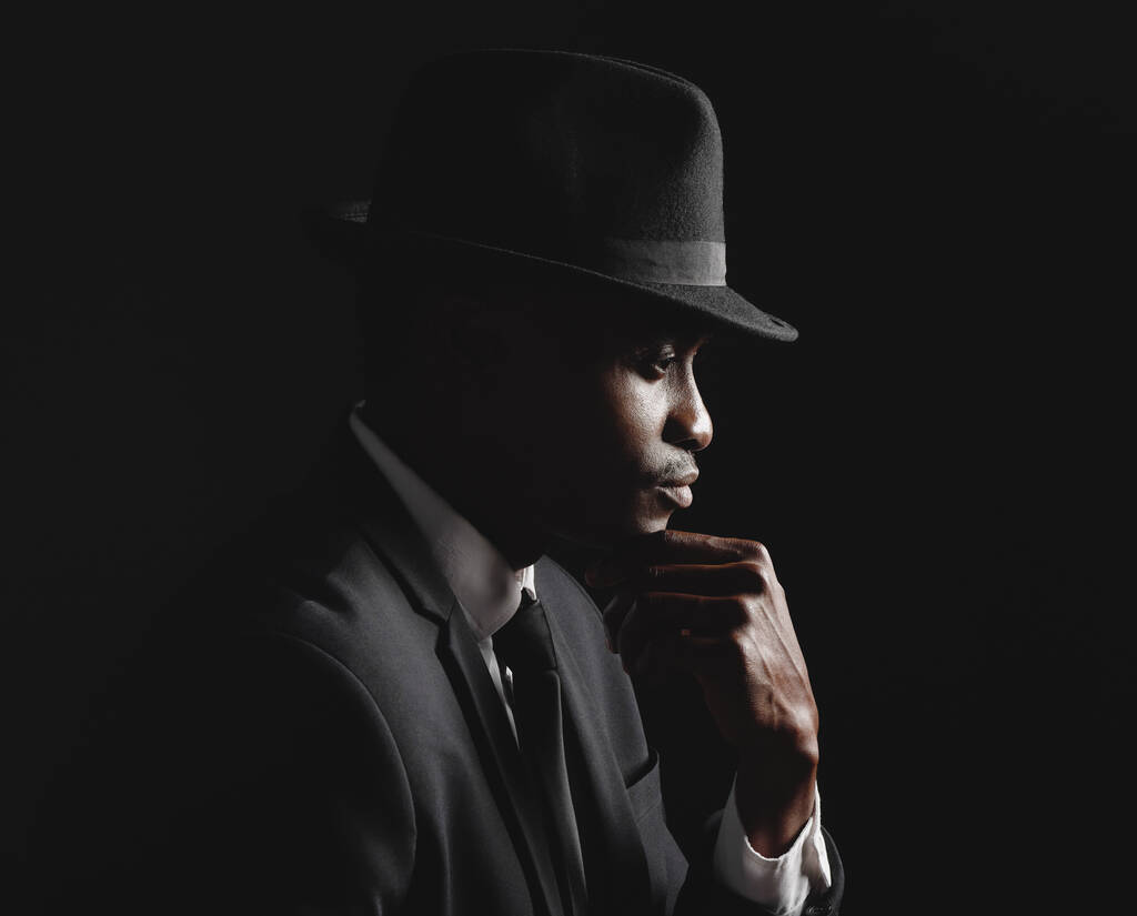 Αφροαμερικάνος που ποζάρει σε μαύρο φόντο φορώντας κοστούμι και καπέλο φαίνεται προσεκτικός και λυπημένος.Θλιμμένο άτομο που υποφέρει από κατάθλιψη και άγχος ενώ πενθεί για την απώλεια.Vintage hitman στη μαφία - Φωτογραφία, εικόνα
