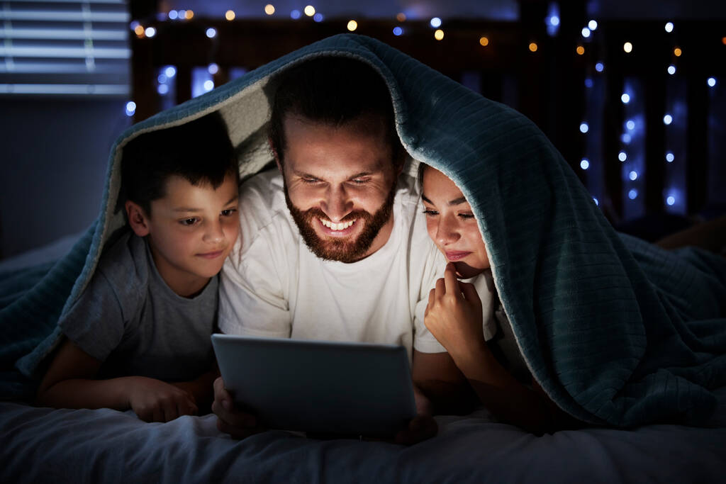 Happy caucasian family single dad with two children using digital tablet lying under blanket in the dark at night with their faces illuminated by device screen light. Father reading online story or - Photo, Image