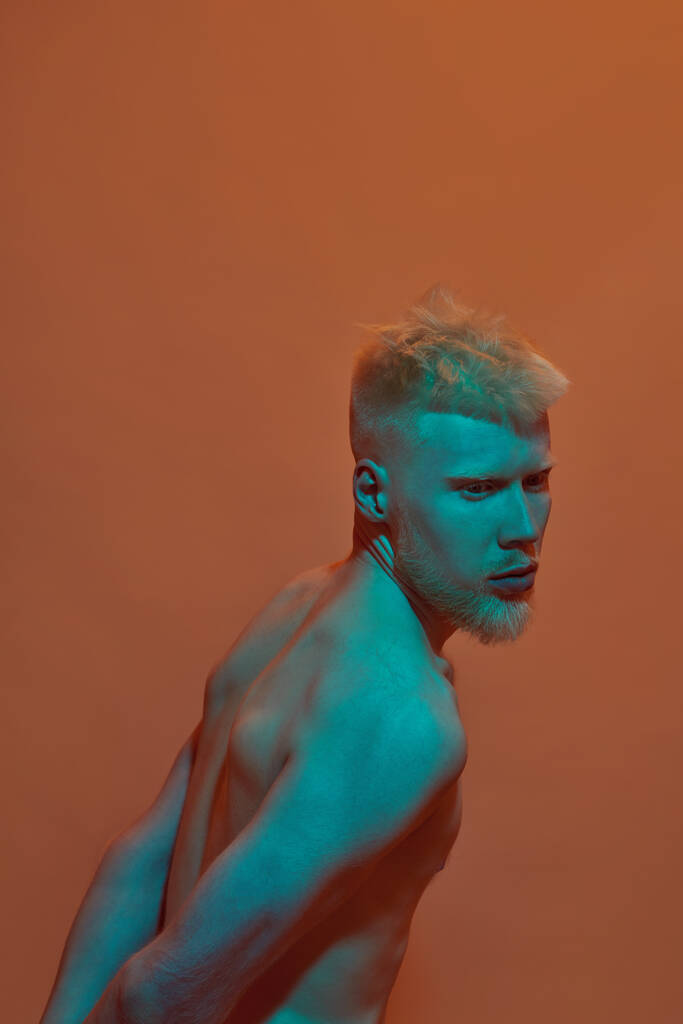 Pensativo chico albino caucásico posando y mirando hacia otro lado. Joven hombre rubio barbudo moderno de generación Z con torso desnudo. Aislado sobre fondo naranja. Sesión de estudio. Copiar espacio - Foto, imagen