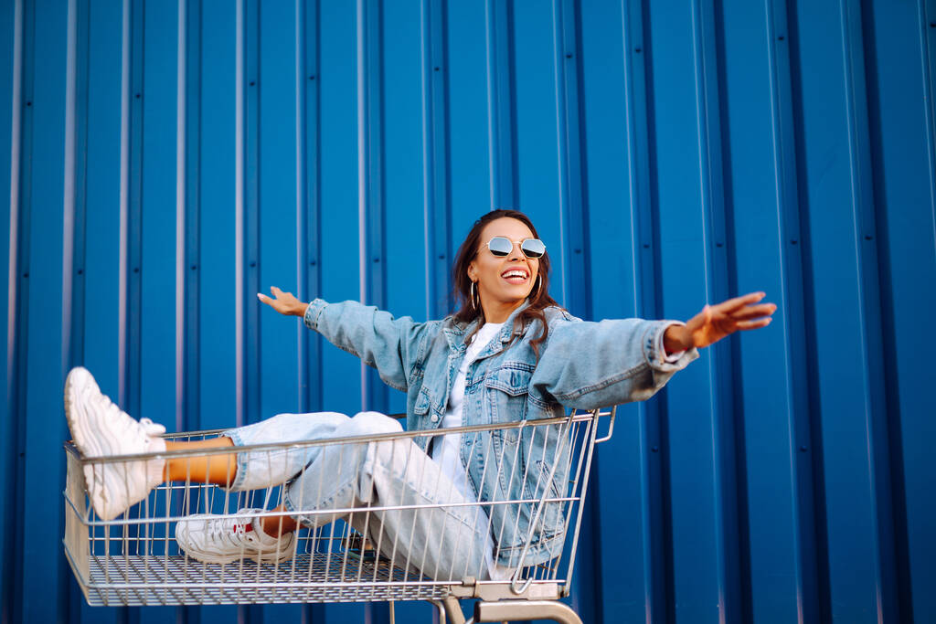 Alışveriş arabalı mutlu kadın renk duvarının yanında eğleniyor. Tüketim, satış, alım, alışveriş, yaşam tarzı konsepti. - Fotoğraf, Görsel