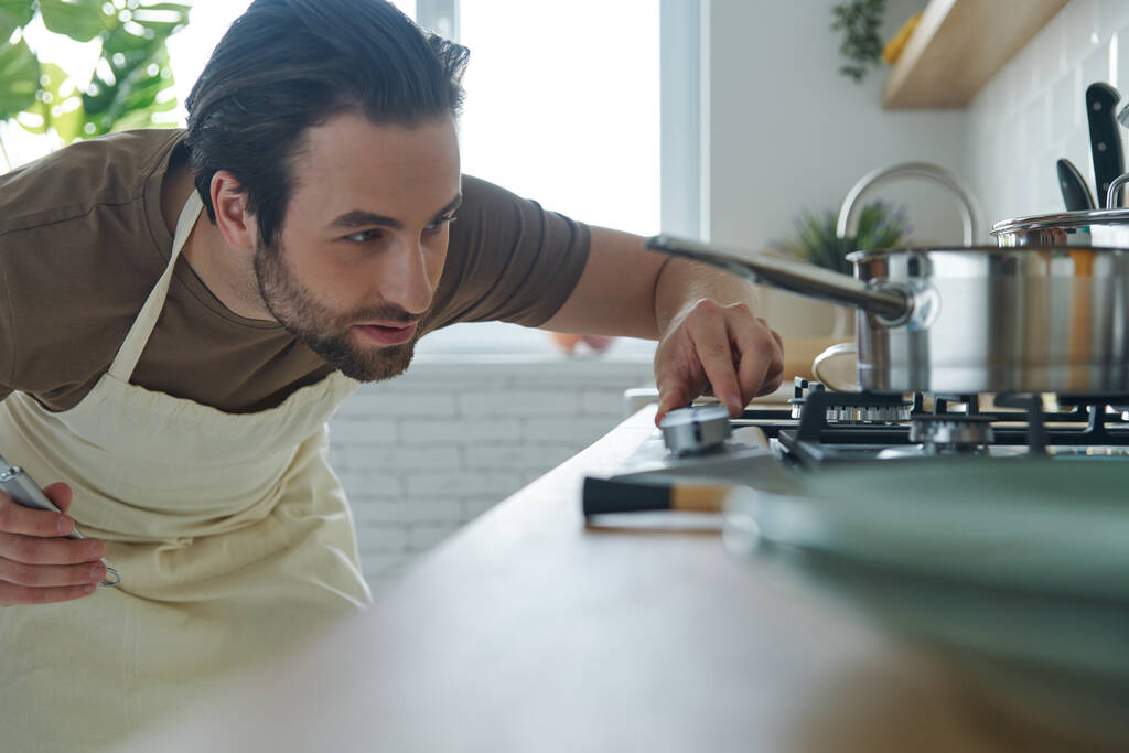 Уверенный молодой человек включил плиту, стоя на кухне - Фото, изображение