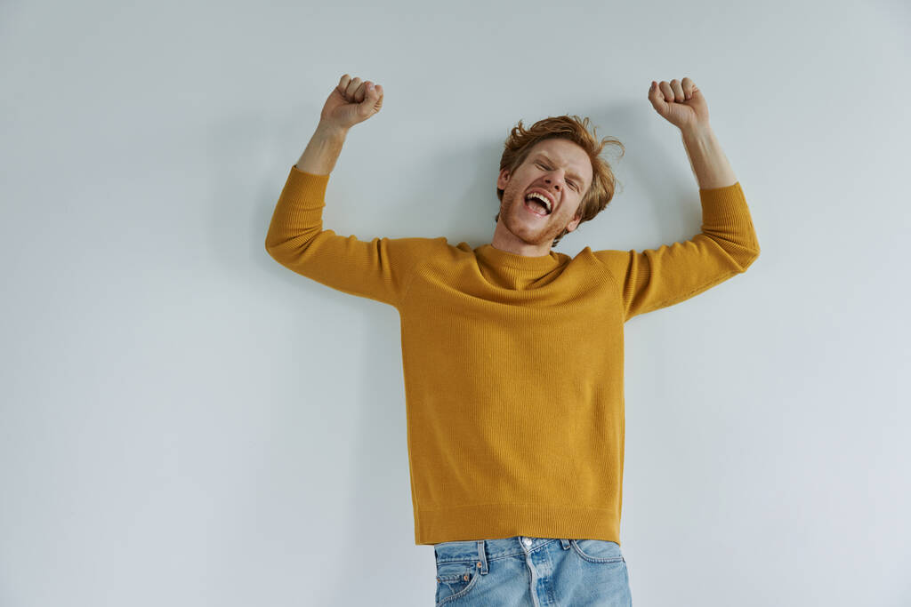 Ευτυχισμένος νεαρός κοκκινομάλλης που κρατάει ψηλά τα χέρια και χαμογελά ενώ στέκεται στον τοίχο. - Φωτογραφία, εικόνα