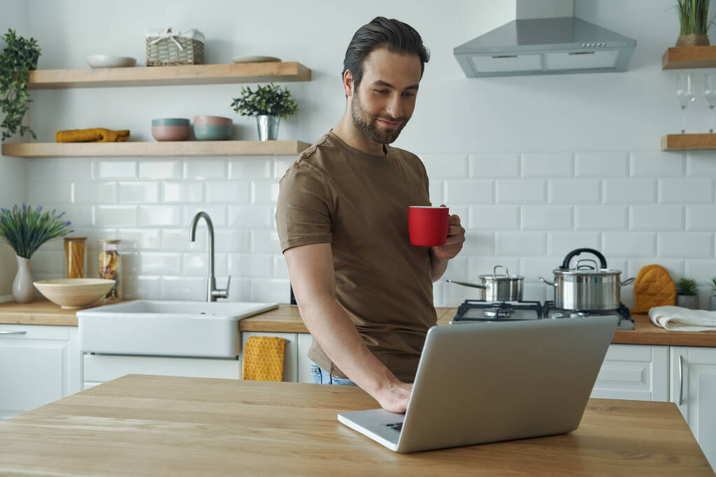 Beau jeune homme utilisant un ordinateur portable et profitant du café tout en se tenant à l'îlot de cuisine - Photo, image