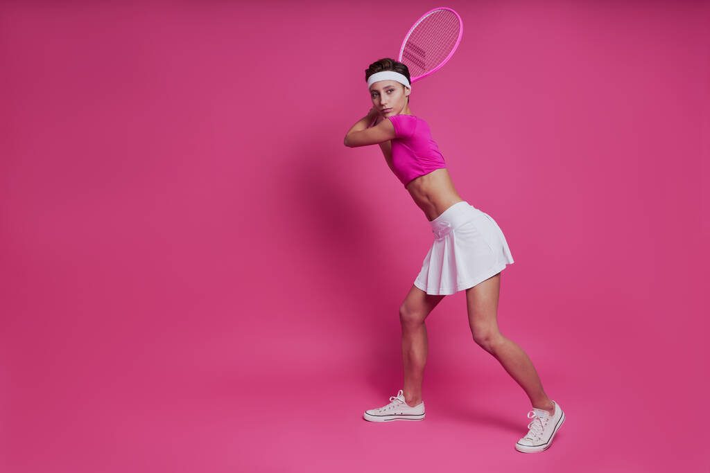 Kendine güvenen genç bir kadın pembe arka planda dururken elinde tenis raketi tutuyor. - Fotoğraf, Görsel