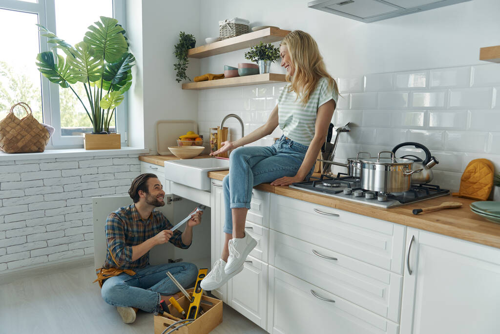 Уверенный молодой человек чинит раковину, пока женщина сидит на кухонном столе и улыбается - Фото, изображение