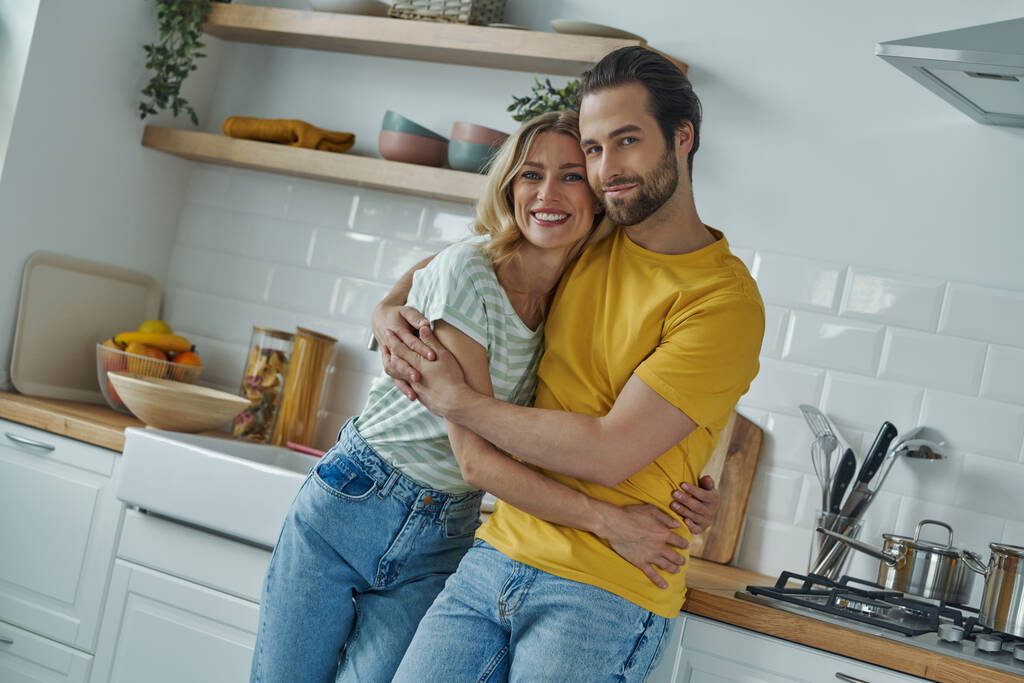 Gyönyörű fiatal pár ölelkezik és a kamerába néz, miközben a konyhában állnak. - Fotó, kép