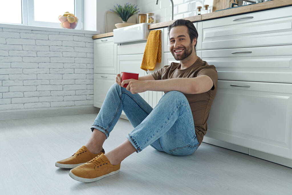 Happy young man enjoying coffee while sitting on the floor and leaning at the kitchen counter - Photo, Image