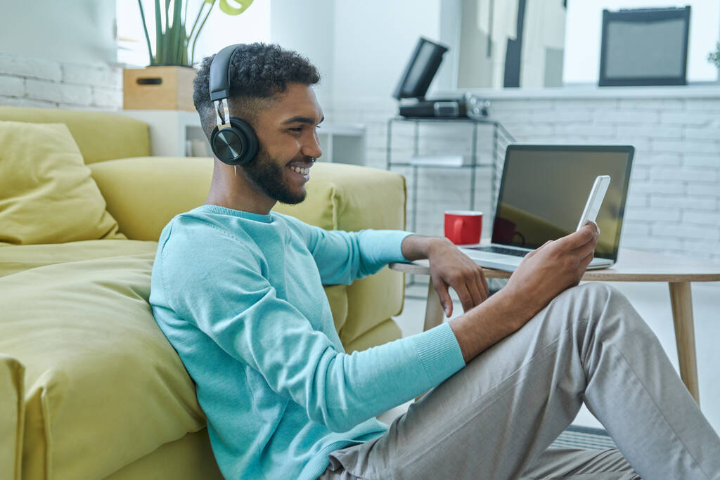 Joyful African man in headphones enjoying music while sitting on the floor at home - Photo, Image