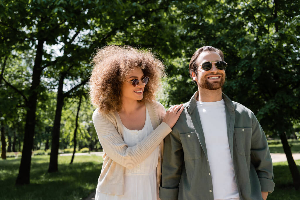 curly woman and happy man in stylish sunglasses walking in park - Photo, Image