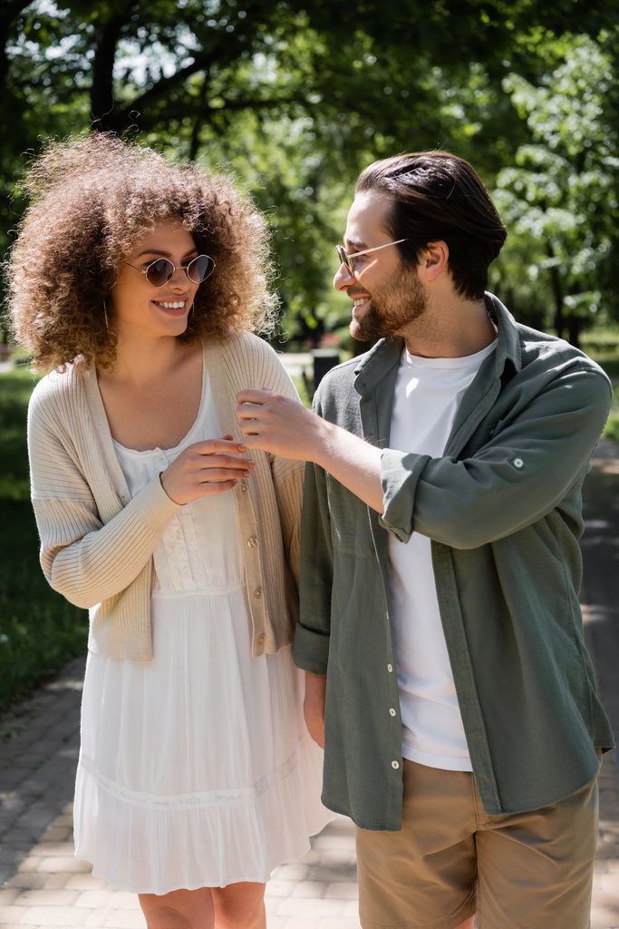 curly woman and cheerful man in stylish sunglasses walking in park - Photo, Image