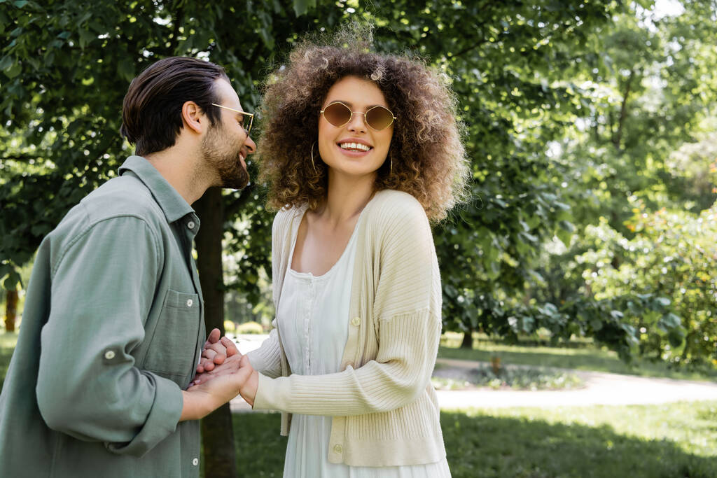 χαρούμενη γυναίκα κρατώντας τα χέρια με τον χαρούμενο άνδρα σε κομψά γυαλιά ηλίου στο πάρκο - Φωτογραφία, εικόνα