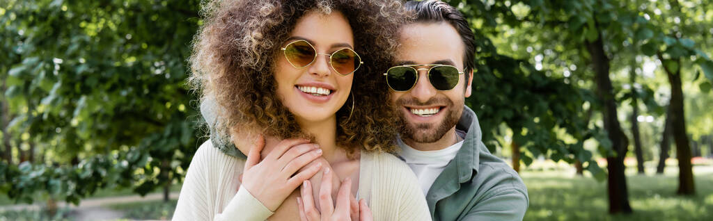 happy man in stylish sunglasses hugging curly girlfriend in park, banner - Foto, Imagem