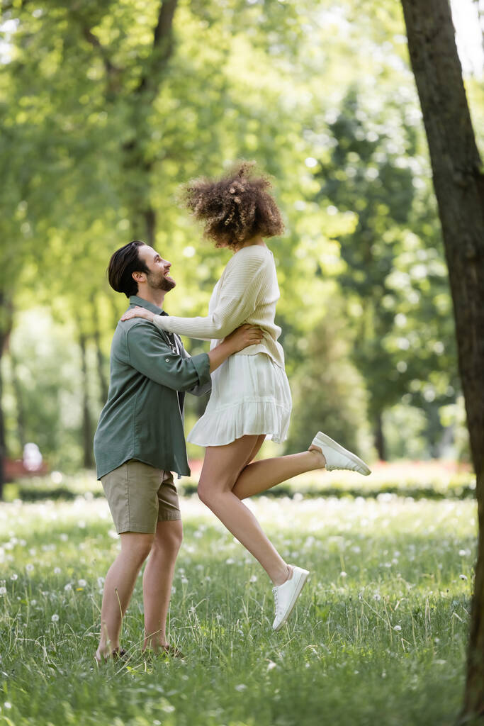 full length of happy young man lifting curly girlfriend in summer dress in green park  - 写真・画像