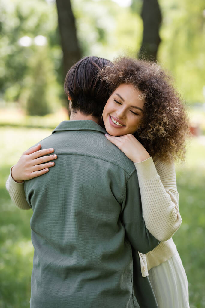 cheerful and curly woman in cardigan hugging boyfriend in green park - Foto, immagini