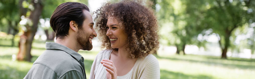 happy and curly woman in cardigan looking at cheerful boyfriend in green park, banner - 写真・画像