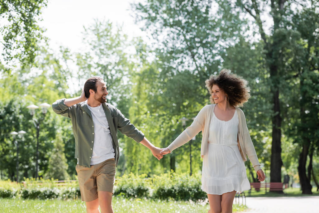 potěšený mladý pár držící se za ruce při procházce spolu v letním parku  - Fotografie, Obrázek