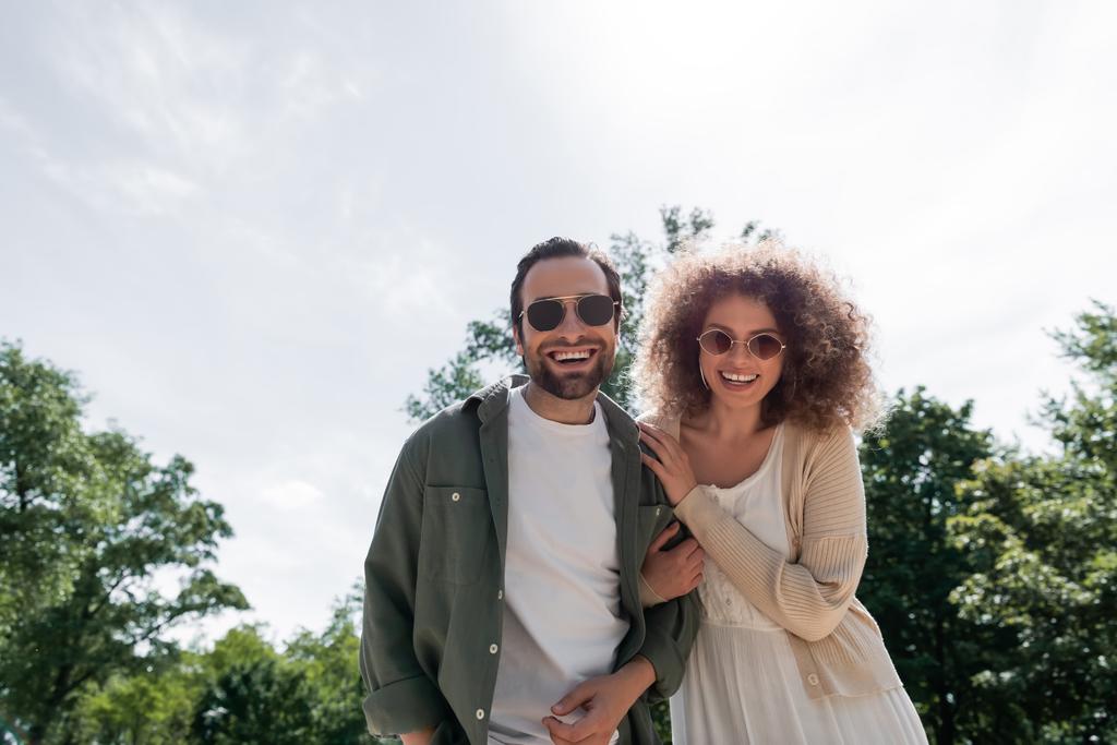 cheerful man and happy curly woman in trendy sunglasses smiling in park  - 写真・画像