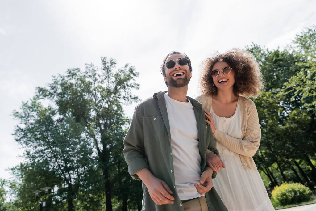 joyful man and happy curly woman in trendy sunglasses smiling in park  - 写真・画像