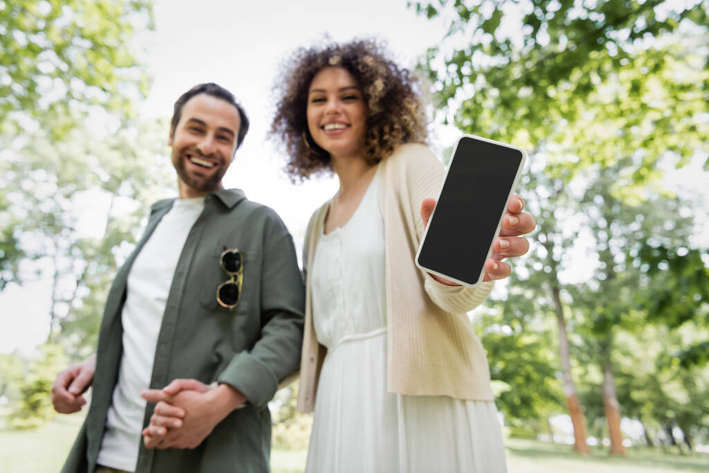 vista de ángulo bajo de la mujer rizada feliz sosteniendo teléfono inteligente con pantalla en blanco cerca de novio sonriente en el parque - Foto, Imagen