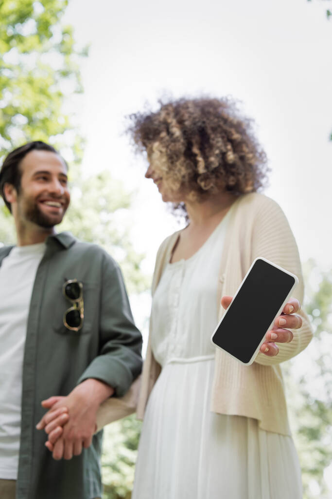 χαμηλή γωνία άποψη του σγουρά γυναίκα κρατώντας smartphone με λευκή οθόνη κοντά χαμογελαστό φίλο στο πάρκο - Φωτογραφία, εικόνα