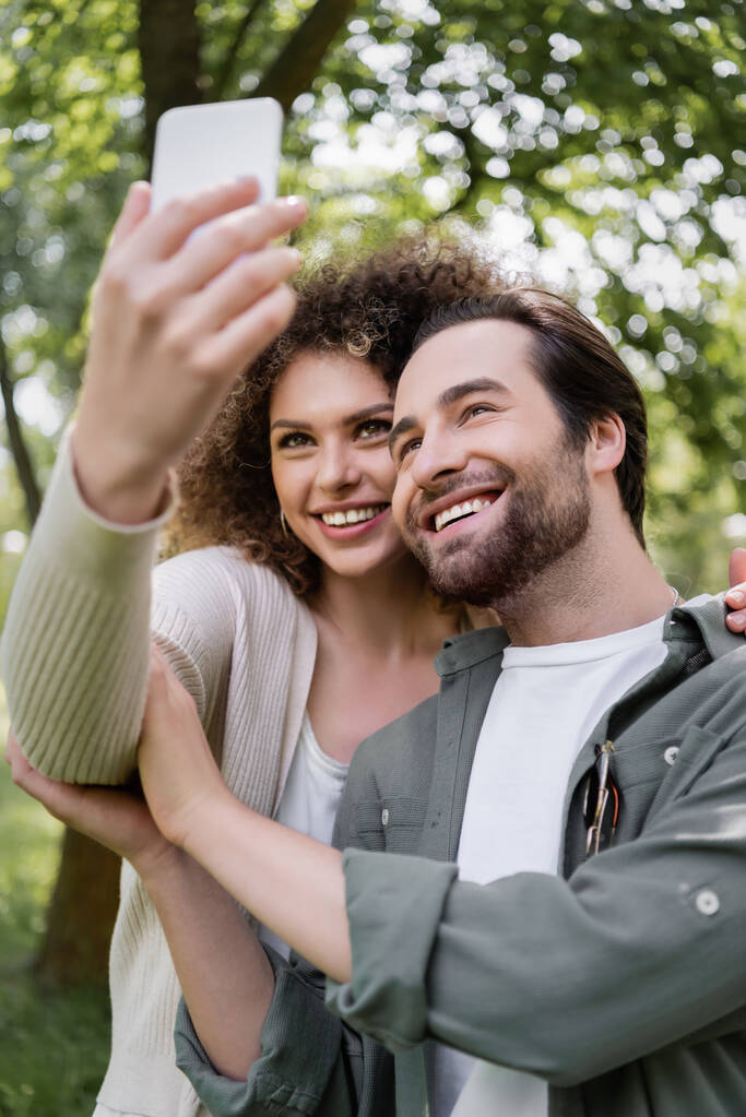 χαρούμενο νεαρό ζευγάρι που παίρνει selfie σε θολή smartphone στο πράσινο καλοκαιρινό πάρκο  - Φωτογραφία, εικόνα
