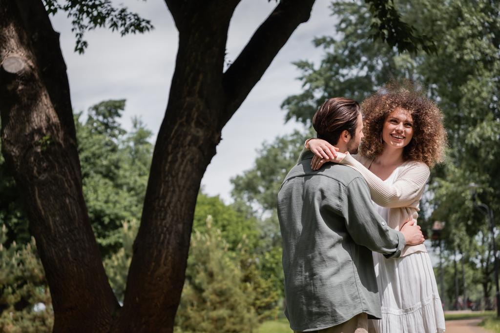 ευτυχισμένη σγουρά γυναίκα αγκαλιάζει γενειοφόρο άνδρα κατά τη διάρκεια του ραντεβού στο πάρκο καλοκαίρι  - Φωτογραφία, εικόνα