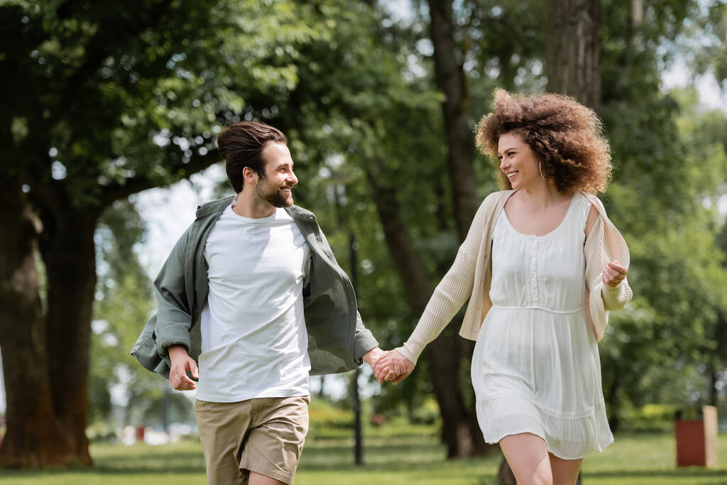 счастливая пара в летней одежде, держась за руки в зеленом парке  - Фото, изображение