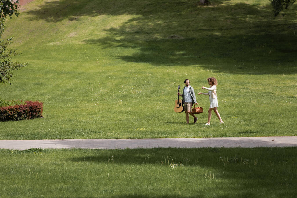 πλήρες μήκος του ανθρώπου σε καλοκαιρινά ρούχα που μεταφέρουν κιθάρα και ψάθινο καλάθι κοντά στην ευτυχισμένη φίλη gesturing στο πράσινο πάρκο  - Φωτογραφία, εικόνα
