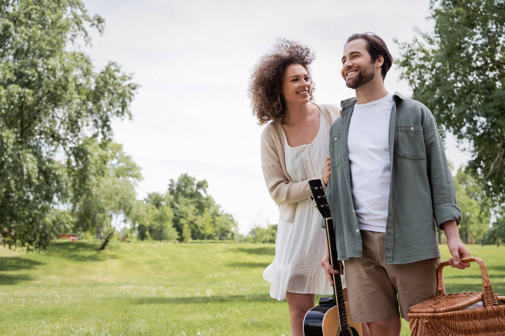man in summer clothes carrying guitar and wicker basket near happy girlfriend in green park  - Fotografie, Obrázek