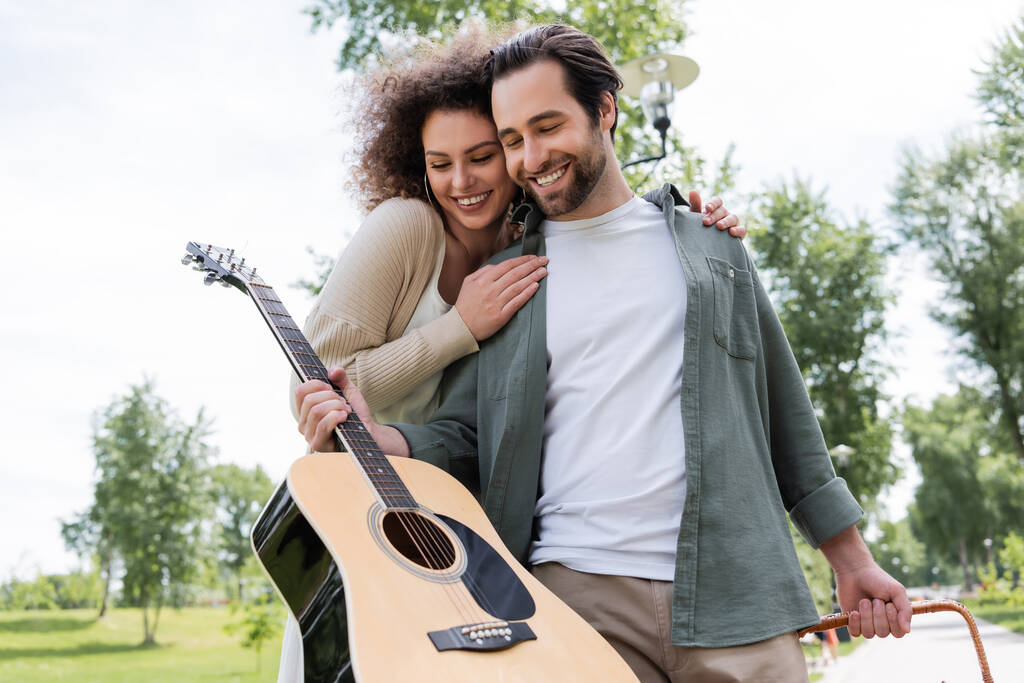 uśmiechnięty mężczyzna w letnich ubraniach trzyma gitarę w pobliżu szczęśliwej dziewczyny w zielonym parku  - Zdjęcie, obraz