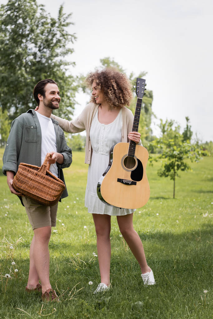plná délka kudrnaté ženy drží akustickou kytaru v blízkosti šťastný přítel s proutěný koš v zeleném parku - Fotografie, Obrázek